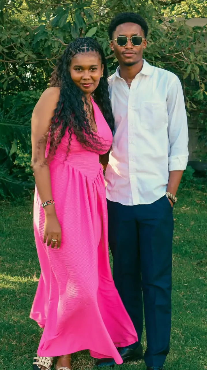 A couple dressed for celebration standing close together in a sunlit garden, she in pink, he in white