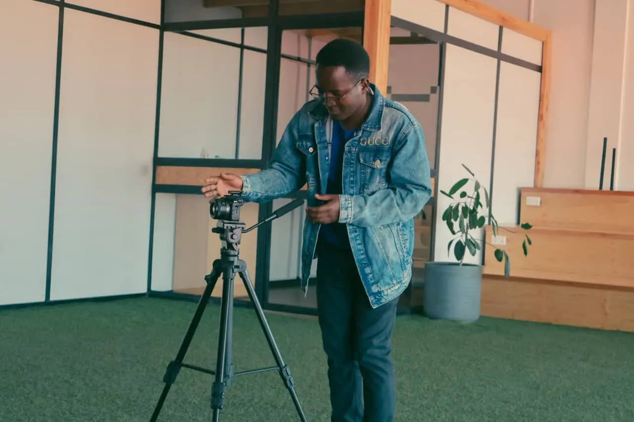 Ian Asena in a denim jacket adjusting a camera on a tripod inside a bright studio space