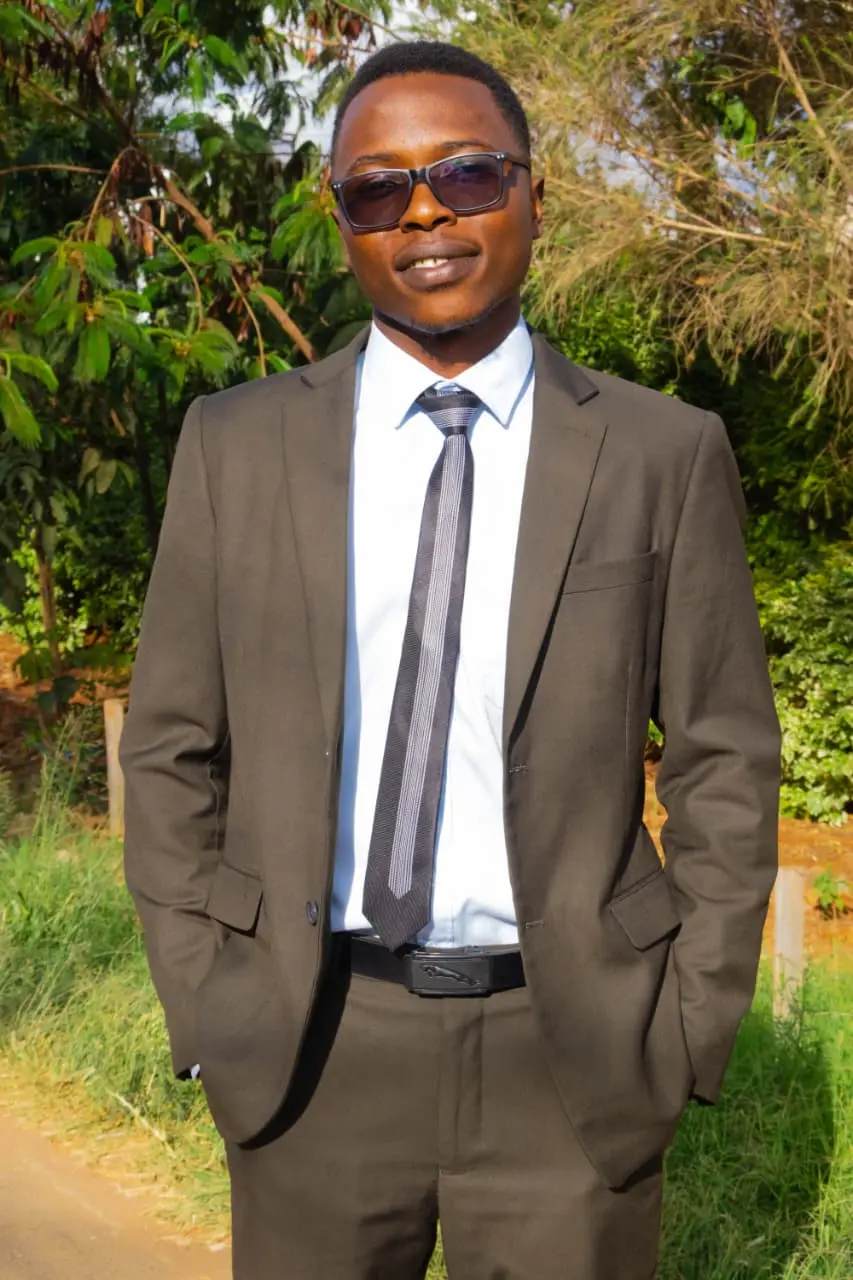 A graduate-to-be in a sharp brown suit and sunglasses, photographed among green trees